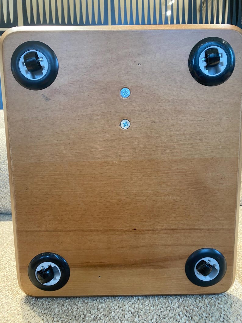 Puede incluir: Base de caja de madera marr&oacute;n claro con cuatro ruedas negras, una en cada esquina. Las ruedas tienen detalles blancos y negros. Dos peque&ntilde;os tornillos azul claro son visibles en el centro de la base.
