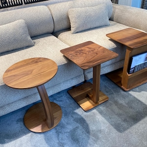 Peut inclure: Trois tables d'appoint en bois de différentes formes. L'une a un plateau ovale et un pied incurvé, une autre a un plateau carré, et la troisième a un plateau carré avec un porte-revues. Toutes sont en bois brun clair.