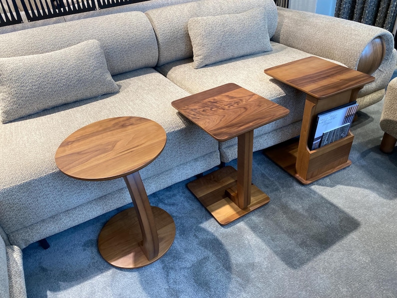Puede incluir: Tres mesas auxiliares de madera con diferentes dise&ntilde;os. Una tiene una tapa ovalada y una pata curva, otra tiene una tapa cuadrada, y la tercera tiene una tapa cuadrada con un revistero. Todas las mesas son de madera marr&oacute;n claro.