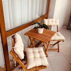 May include: A small outdoor patio set with a square wooden table and two folding chairs. Each chair has a cream-colored cushion. A potted plant sits on the table. The set is made of light brown wood.
