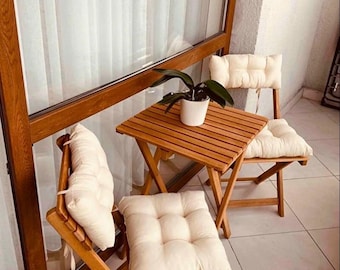 Puede incluir: Un pequeño juego de patio al aire libre con una mesa cuadrada de madera y dos sillas plegables. Cada silla tiene un cojín de color crema. Una planta en maceta se encuentra sobre la mesa. El conjunto es de madera marrón claro.
