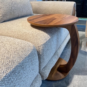 Puede incluir: Una mesa auxiliar de madera con una tapa redonda y un soporte curvo. La mesa es de madera de color marr&oacute;n claro y est&aacute; colocada junto a un sof&aacute; tapizado de color gris claro. El dise&ntilde;o de la mesa es moderno y minimalista.