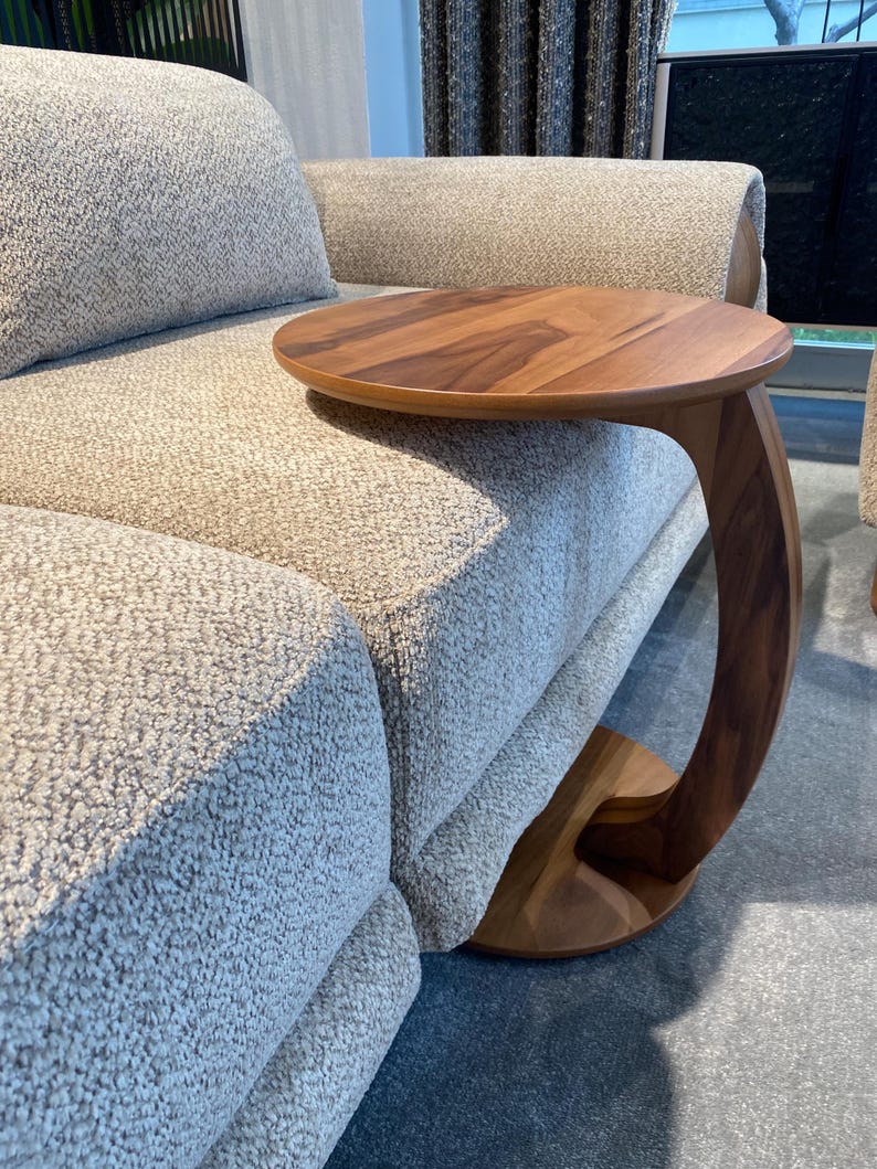Puede incluir: Una mesa auxiliar de madera con una tapa redonda y un soporte curvo. La mesa es de madera de color marr&oacute;n claro y est&aacute; colocada junto a un sof&aacute; tapizado de color gris claro. El dise&ntilde;o de la mesa es moderno y minimalista.