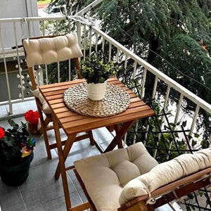 Puede incluir: Un pequeño juego de bistró de madera con dos sillas plegables y una mesa cuadrada. Las sillas tienen cojines color crema. Una planta en maceta se encuentra sobre la mesa, que tiene un salvamanteles tejido. El balcón tiene una barandilla blanca y vistas a árboles.