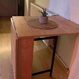 May include: A wooden folding table with a black metal frame. The table is set with a woven placemat and a small potted plant with purple flowers.