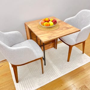 Peut inclure: Une table pliante en bois avec une finition en bois clair et des pieds en métal noir. Deux chaises blanches avec des pieds en bois sont placées de chaque côté de la table. Un bol de fruits est posé sur la table.