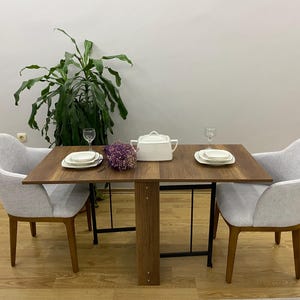 Puede incluir: Una mesa de comedor de madera con dos sillas, puesta para dos con platos y vasos blancos. La mesa tiene una hoja abatible y está hecha de madera oscura. Las sillas están tapizadas en tela gris claro.