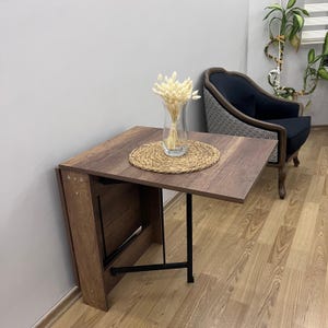 May include: A brown wooden folding table with black metal legs. The table is set with a woven straw placemat and a clear glass vase with dried white flowers.
