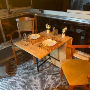 May include: A small wooden table with a black metal frame, set for two with white plates, woven placemats, and clear glasses. The table is set against a backdrop of a window with a view of a city.