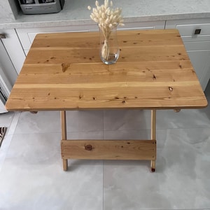 Puede incluir: Una mesa plegable de madera clara con un acabado natural. La mesa tiene un diseño sencillo con una sola tabla para un estante debajo. Un jarrón con flores blancas secas se encuentra sobre la mesa.
