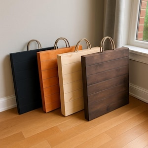May include: Four wooden serving trays in black, orange, natural, and dark brown. Each tray has a rectangular shape with horizontal wooden slats and a rope handle. The trays are leaning against a wall and are displayed on a wooden floor.