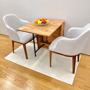 May include: A small wooden table with a drop-leaf extension, two white chairs, and a bowl of fruit on the table.