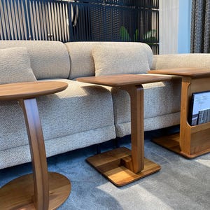 May include: Three wooden side tables in varying designs. One has a round top, another a square top, and the third has a magazine rack. All tables are made of light brown wood and have a modern aesthetic. The tables are placed in front of a beige sofa.