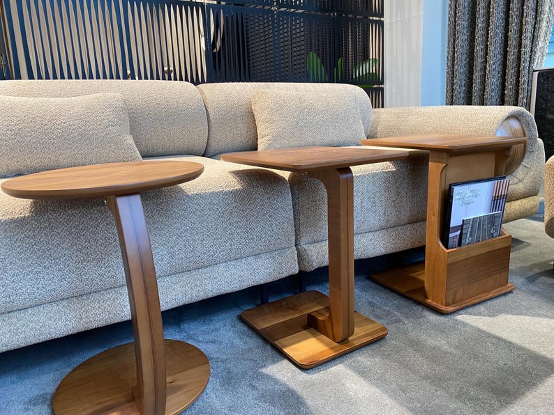 Puede incluir: Tres mesas auxiliares de madera de diferentes dise&ntilde;os. Una tiene una tapa redonda, otra una tapa cuadrada y la tercera tiene un revistero. Todas las mesas son de madera marr&oacute;n claro y tienen una est&eacute;tica moderna. Las mesas est&aacute;n colocadas frente a un sof&aacute; beige.
