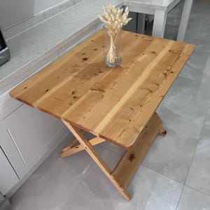 May include: A light wood folding table with a natural finish. The table has a simple, rustic design and is perfect for small spaces. A vase with dried flowers sits on the table.