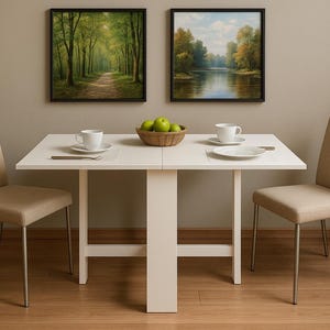 May include: A white dining table with two matching chairs. Two framed landscape paintings hang above the table. A bowl of green apples, two cups, and plates are on the table. The chairs have beige upholstery and metal legs.