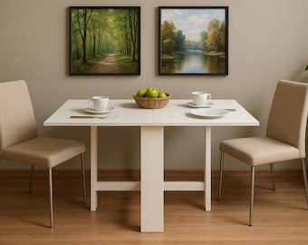 May include: A white dining table with two matching chairs. Two framed landscape paintings hang above the table. A bowl of green apples, two cups, and plates are on the table. The chairs have beige upholstery and metal legs.