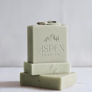 Puede incluir: Tres barras de jabón verde apiladas una encima de la otra. La barra superior tiene las palabras "Aspen Soap Co." en relieve en la parte delantera. El logotipo presenta una cordillera y pinos.