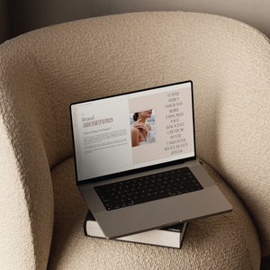 May include: A laptop computer open to a webpage with the text "Brand Archetypes" and a list of brand archetypes including "Lover", "Hero", "Magician", "Rebel", "Explorer", "Sage", "Innocent", "Creator", "Ruler", "Caregiver", "Regular Guy", and "Jester". The laptop is sitting on a stack of books on a beige armchair.