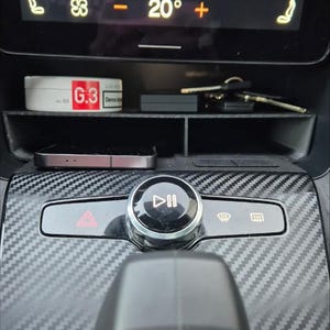 May include: A black car interior with a carbon fibre pattern on the dashboard. The dashboard has a control panel with a play/pause button, a hazard light button, and two other buttons. The temperature control display shows 20 degrees.
