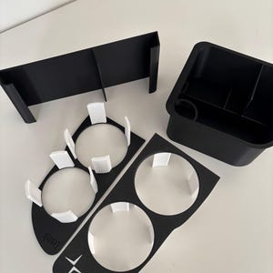 May include: A set of black and white car interior accessories. The image shows a rectangular divider, a storage bin, and two cup holder inserts with white details. The cup holder inserts have the text "Lenteh" and a white star-like symbol.
