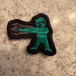 May include: A green and black embroidered patch of a cartoon character shooting a laser beam from their hand.