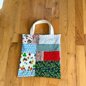 May include: A patchwork tote bag with a white canvas handle. The bag is made of different fabrics with various patterns, including floral, gingham, and geometric designs. The bag is a light blue, red, green, and white color.