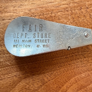 May include: A vintage metal key tag from the Fair Dept. Store, with the store's address in Weirton, W. VA. The tag has a teardrop shape with engraved text and a decorative design on the end. The tag is silver in color.