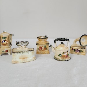 May include: Five miniature ceramic figurines of household items, including a stove, iron, teapot, and gramophone. Each figurine is decorated with a floral pattern and painted in shades of cream, brown, and green.