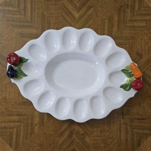 Peut inclure: Plat à servir en céramique blanche avec un bord festonné et plusieurs cavités ovales. Le plat présente des poignées décoratives avec des légumes peints, dont une tomate rouge, une aubergine violette et une carotte orange.