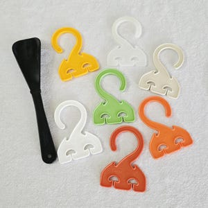 May include: A black shoe horn and several colorful plastic shoe hangers. The hangers are in yellow, white, green, orange, and off-white. The hangers have a unique design with a hook and a mushroom-shaped base.