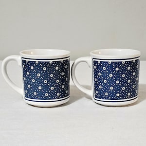 May include: A pair of white ceramic mugs featuring a dark blue rectangular panel on the front. The blue panel is decorated with a repeating pattern of small white flowers and thin white lines, framed by a solid white border on the mug surface.