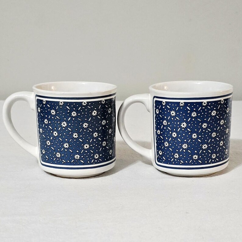 May include: A pair of white ceramic mugs featuring a dark blue rectangular panel on the front. The blue panel is decorated with a repeating pattern of small white flowers and thin white lines, framed by a solid white border on the mug surface.