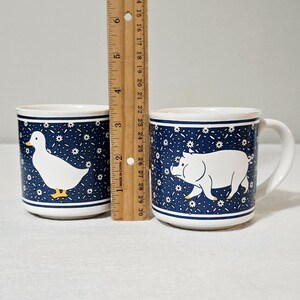 May include: Two white ceramic mugs with a dark blue background featuring a white daisy pattern. One mug displays a white duck with a yellow beak and feet, while the other features a white pig. A wooden ruler shows the mugs are 3.5 inches tall.