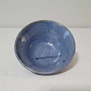 May include: A small, handmade ceramic bowl in a muted blue hue with a textured, matte finish. The bowl has an irregular shape and a brown rim. The interior shows subtle colour variations. The bowl is suitable for serving small portions.