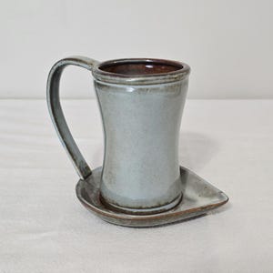 May include: A handmade ceramic mug and saucer set. The mug is light blue with a curved handle and a brown rim. The saucer is a matching light blue with a brown rim. The set is designed for coffee or tea.