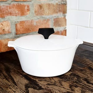 May include: A white ceramic casserole dish with a matching lid and integrated side handles. The lid features a distinct, black, geometric knob handle centered on top. The vessel has a smooth, glossy finish and a wide, circular base design.