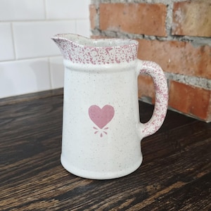 May include: White ceramic pitcher with a pink heart design and speckled accents. The handle and rim feature a pink speckled pattern. The pitcher is on a dark wooden surface.