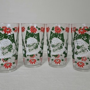 May include: Four clear drinking glasses with a festive design. Each glass features a green bow, white bell, red poinsettias, and the text "Happy New Year". The glasses are perfect for holiday celebrations.