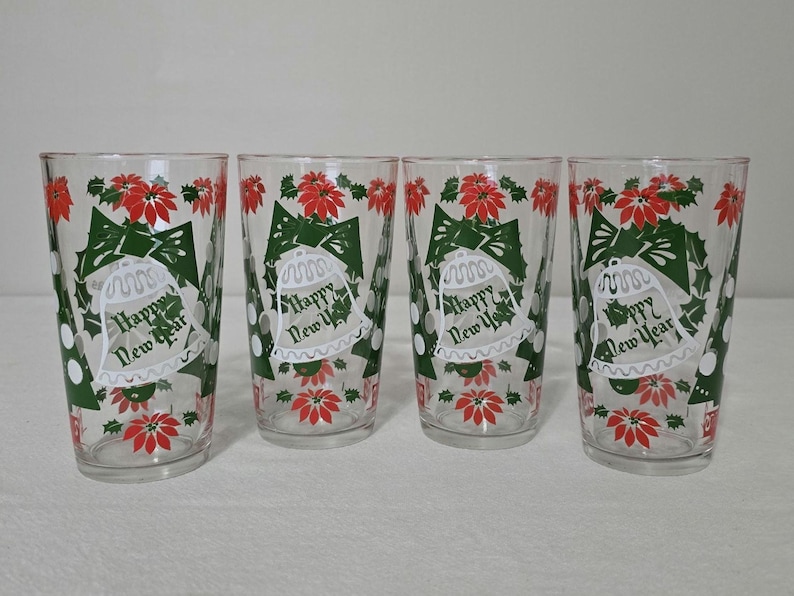 May include: Four clear drinking glasses with a festive design. Each glass features a green bow, white bell, red poinsettias, and the text "Happy New Year". The glasses are perfect for holiday celebrations.