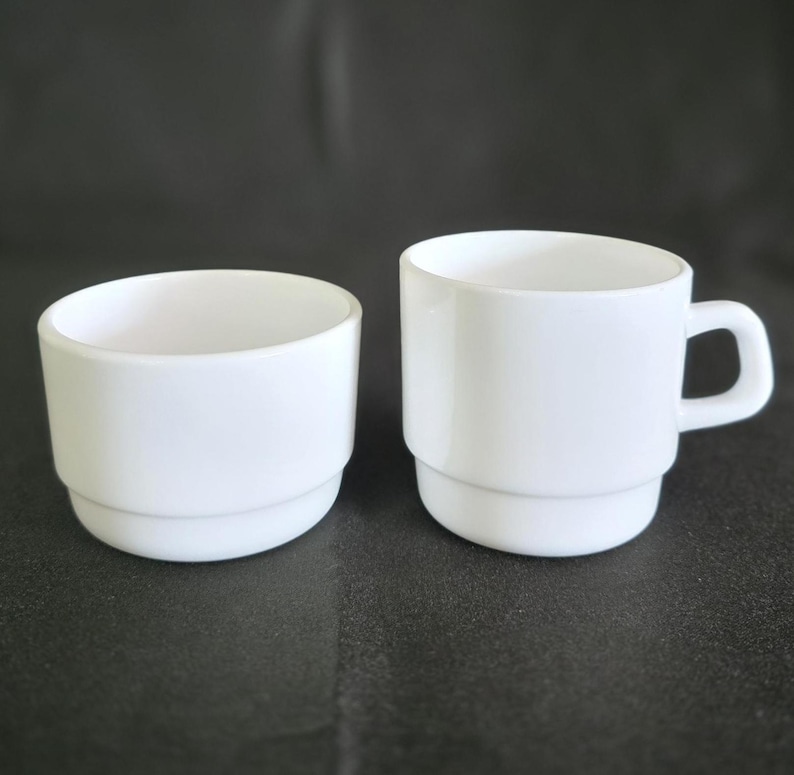 May include: Two white ceramic cups, one with a handle and the other without. The cup with the handle is stacked with two layers.
