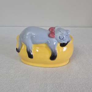 May include: A ceramic figurine of a gray cat with a pink bow sleeping on a yellow oval-shaped bed.