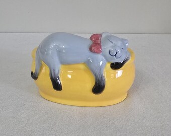 May include: A ceramic figurine of a grey cat with a pink bow sleeping on a yellow oval-shaped bed.