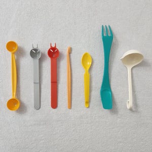 May include: A set of seven kitchen utensils, including a yellow egg separator, a gray egg slicer, a red egg topper, an orange tool, a yellow spoon, a teal spatula, and a white ladle.