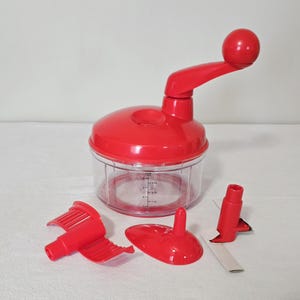 May include: A red manual food chopper with a clear plastic container and a handle. The chopper includes several red accessories, including a blade and a lid. The container has measurement markings in cups and ounces.