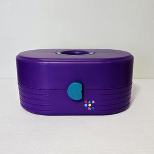 May include: A purple plastic storage case with an oval shape and a teal latch. The top features a recessed handle, and the front displays the word CABOODLES in small, colorful geometric letters. The lower section has horizontal ribbed detailing.