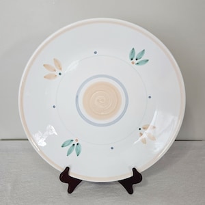 May include: White ceramic plate with a beige rim and floral accents. The plate features a central spiral design in beige and blue, with small blue dots and leaf-like designs in beige and teal. The plate is displayed on a dark brown stand.