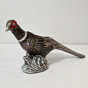 May include: A ceramic pheasant figurine in shades of brown, gray, and green. The bird has a red eye ring and a silver-colored neck band. The pheasant is perched on a decorative base.