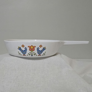 May include: White ceramic skillet with a long handle. The skillet features a colorful floral and bird design in blue, orange, and yellow. The design is centered on the front of the skillet.