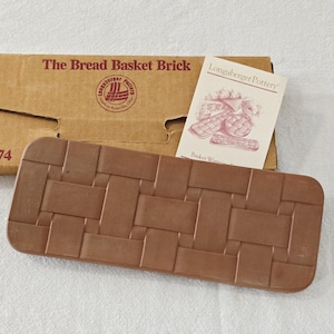 May include: A brown ceramic bread basket brick with a woven basket pattern. The brick is next to a cardboard box with the text "The Bread Basket Brick" and a Longaberger Pottery logo. A small card with basket illustrations is also present.
